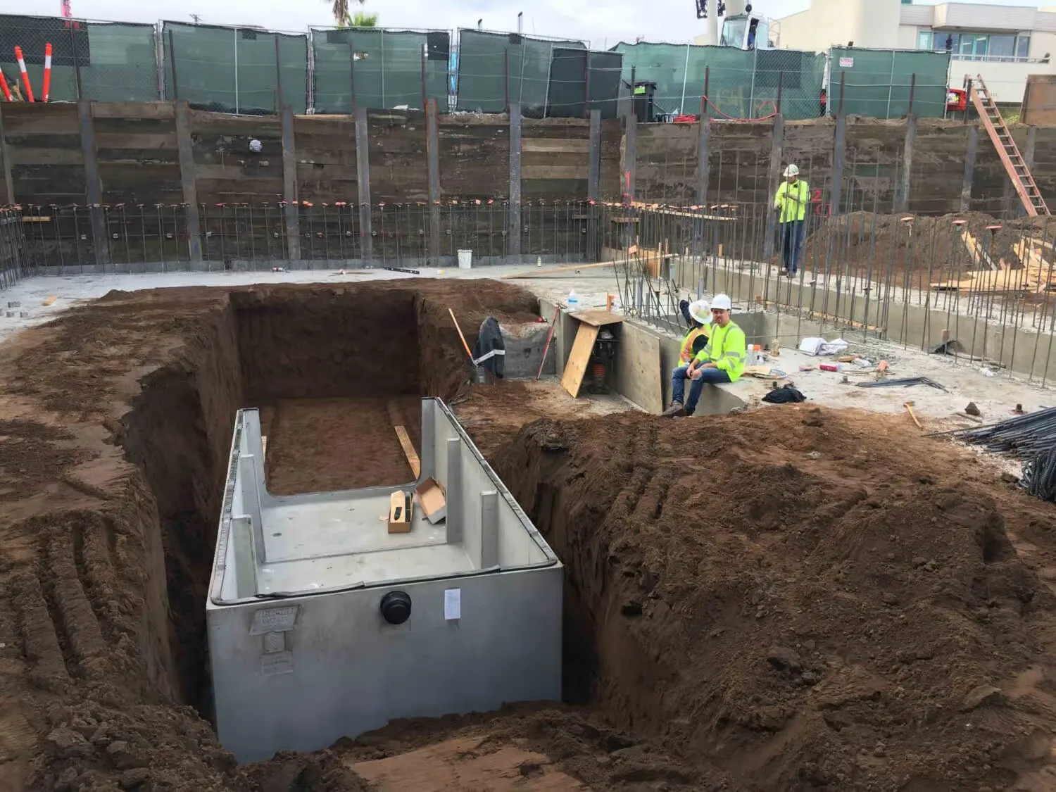Construction site with a large underground concrete tank installation, excavation work, reinforcing steel, and workers wearing safety gear.