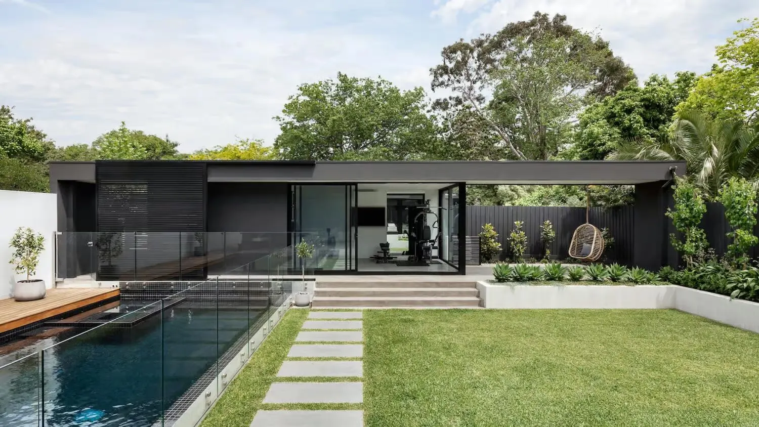 Modern single-story home with a landscaped backyard, glass-fenced swimming pool, stepping stone pathway, covered patio, and lush green lawn surrounded by mature trees.